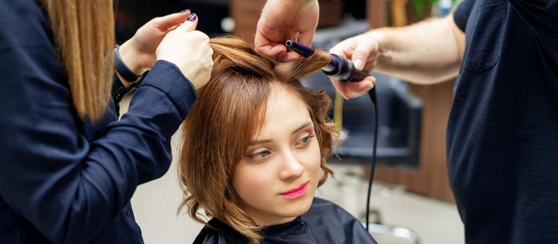 Professional hairdresser twists curls of long light brown hair of woman with curling iron in beauty salon. Hairdressing procedures