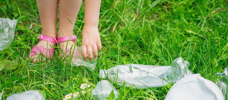 Hand of child cleans green grass from plastic trash in the park