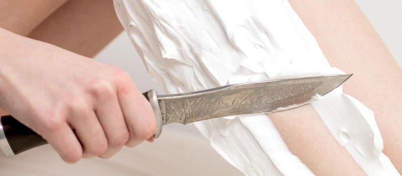 Female beautiful legs of young woman with foam during shaving by knife on white background