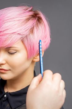 Hairdresser checking short pink hairstyle of young woman on gray background