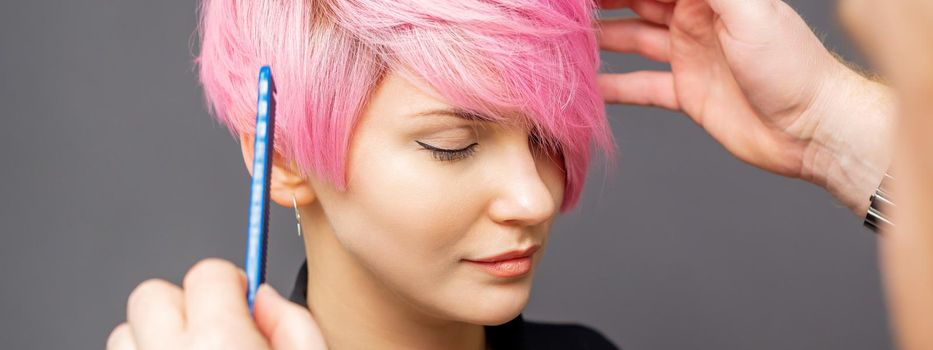 Hairdresser checking short pink hairstyle of young woman on gray background