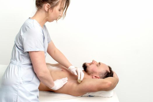 Young man receiving waxing underarm or epilation armpit by young female cosmetologist in beauty salon