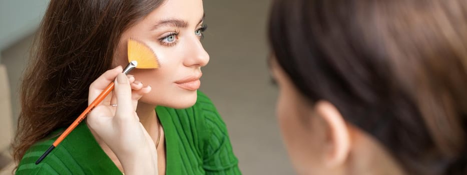 Make up artist applying professional make up of tonal foundation on the face of beautiful young caucasian woman in make up room. Base for make up.