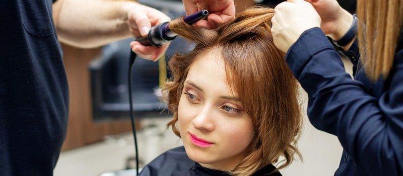 Professional hairdresser twists curls of long light brown hair of woman with curling iron in beauty salon. Hairdressing procedures