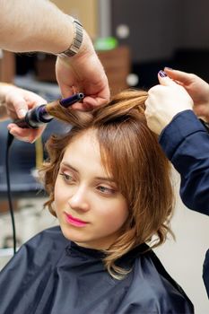 Professional hairdresser twists curls of long light brown hair of woman with curling iron in beauty salon. Hairdressing procedures