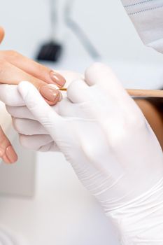 Manicure master in protective gloves applying beige nail polish on female nails in beauty salon