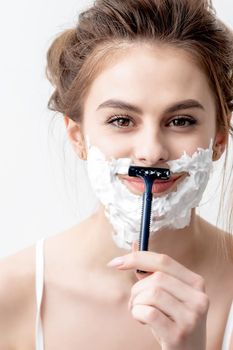 Beautiful young caucasian woman shaving her face by razor on white background. Pretty woman with shaving foam on her face