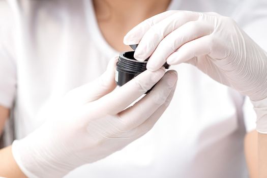 Beautician or manicure master with protective mask holds black jar with cosmetics or manicures product in her hands with white gloves