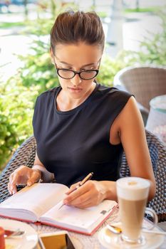 Young caucasian business woman in black dress writes in notebook sitting at table in cafe