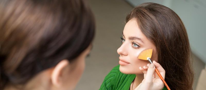 Make up artist applying professional make up of tonal foundation on the face of beautiful young caucasian woman in make up room. Base for make up.