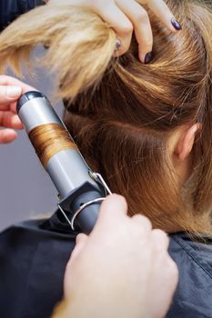 Professional hairdresser twists curls of long light brown hair of woman with curling iron in beauty salon. Hairdressing procedures