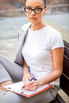 Beautiful young caucasian woman sits on the bench with smartphone and notepad over city street background