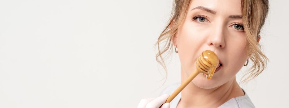 Portrait of beautiful young caucasian woman eating honey with wooden spoon on white background