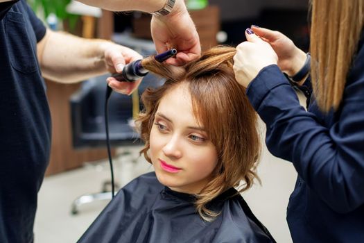 Professional hairdresser twists curls of long light brown hair of woman with curling iron in beauty salon. Hairdressing procedures