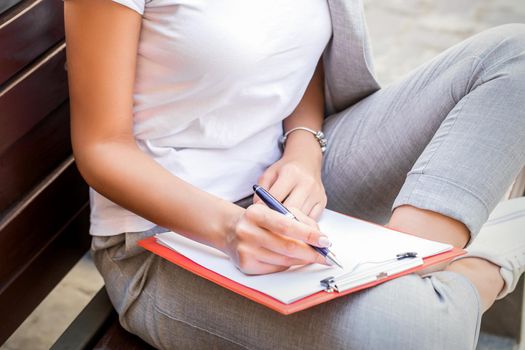 Beautiful young caucasian woman sits on the bench with smartphone and notepad over city street background