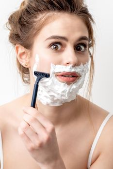 Beautiful young caucasian woman shaving her face by razor on white background. Pretty woman with shaving foam on her face