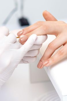 Manicure master in protective gloves applying beige nail polish on female nails in beauty salon
