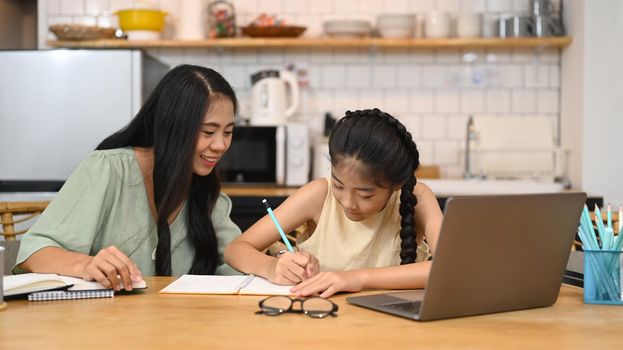 Asian mom helping her sweet daughter doing homework, learning online at virtual class on laptop computer at home.