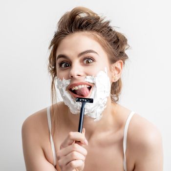 Beautiful young caucasian woman shaving her face by razor on white background. Pretty woman with shaving foam on her face