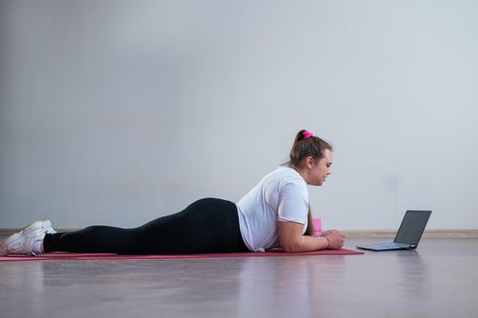 Young Plus Size Woman Stretching At Home Online. Flexible girl practices yoga and watches an online course on a laptop.