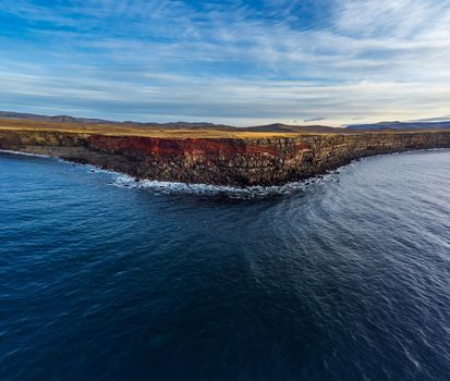 Wide angle view from the ocean of volcanic steep cliffs with red seams in Iceland