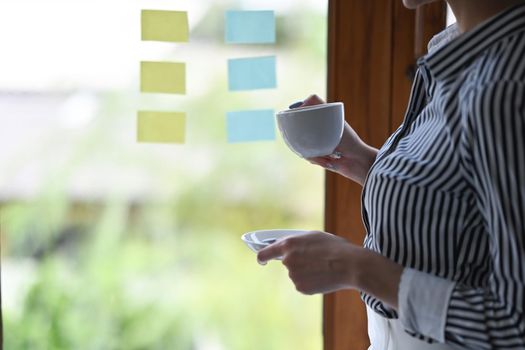 Businesswoman holding coffee cup and reading information on sticky notes at bright office.