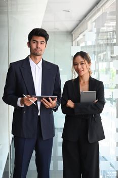 Two professional businesspeople holding digital tablet and looking at camera while standing in office lobby hall.