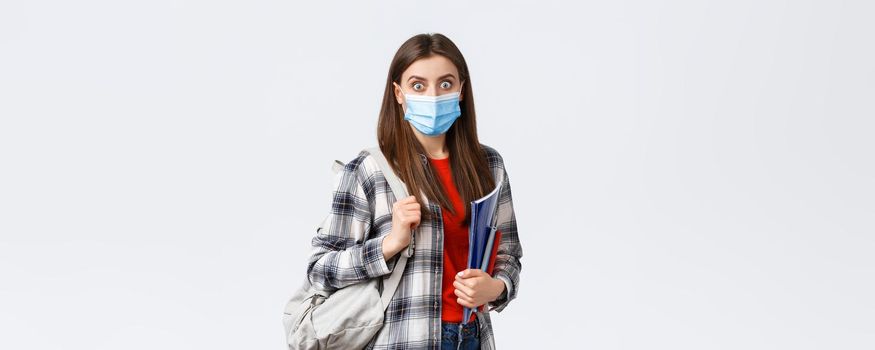 Coronavirus pandemic, covid-19 education, and back to school concept. Shocked and surprised girl in medical mask, student gasping over big news in campus, hold notebooks and backpack.