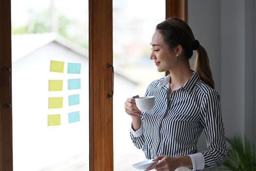 Beautiful female entrepreneur with coffee cup standing near window in office.