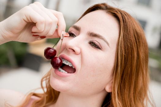 Beautiful young red-haired woman with braces on her teeth eats sweet cherries in the summer outdoors.