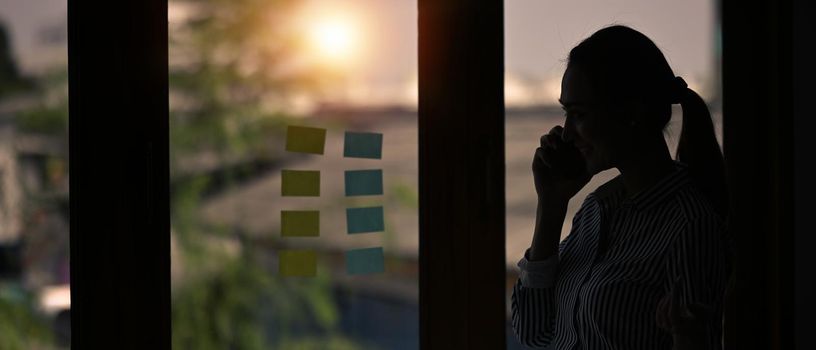 Silhouette of young woman talking on mobile phone while standing near window with view on sunset in city.