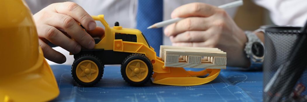 Close-up of yellow miniature toy excavator on blueprint carry wooden blocks. Architect recreates real construction site. Business, building project concept