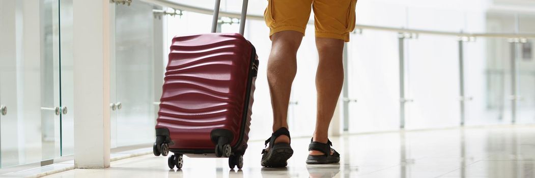 Close-up of young man pulling red suitcase in modern airport terminal. Person going on vacation to hot country. Traveling, airport, departure time concept