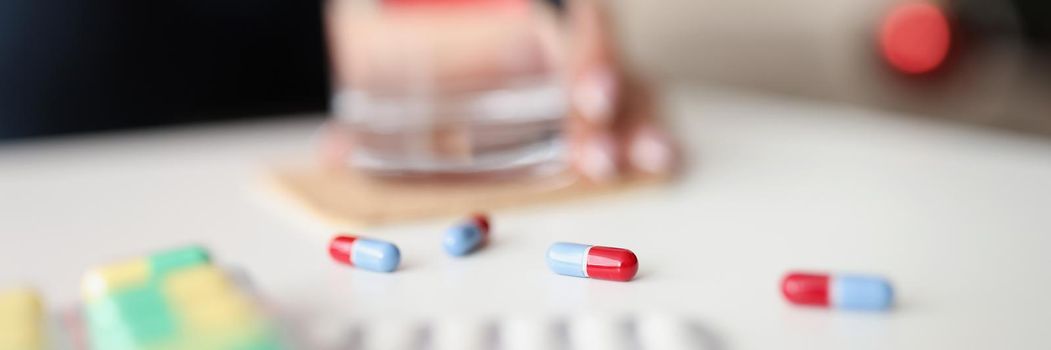 Close-up of jelly capsules scattered on table daily dose of medication for person. Woman hold glass of water on background. Medicine, prescription concept