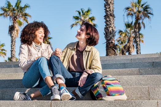 young interracial couple of gay women holding hands laughing sitting on the stairs, concept of sexual freedom and racial diversity