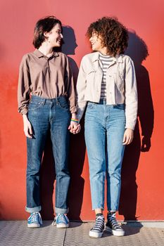 young interracial gay female couple looking at each other holding hands in front of a red wall, concept of sexual freedom and racial diversity, vertical photo
