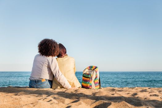 interracial gay female loving couple in front of the sea next to a backpack with the rainbow flag colors, symbol of the struggle for homosexual rights, concept of sexual freedom and racial diversity