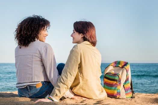 girls in love smiling on the beach next to a backpack with the rainbow flag colors, symbol of the struggle for homosexual rights, concept of sexual freedom and racial diversity