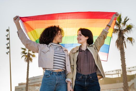 multiracial lesbian couple of smiling women holding hands and raising the rainbow flag, symbol of the struggle for gay rights, concept of sexual freedom and racial diversity