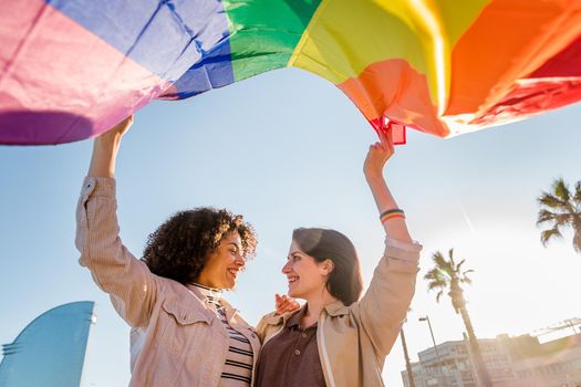 smiling young lesbian couple waves the rainbow flag, symbol of the struggle for gay rights, in Barcelona gay friendly city, concept of sexual freedom and racial diversity