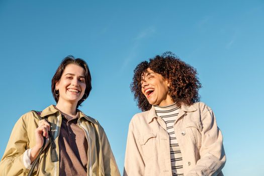 two young women laughing animatedly against a background of blue sky, concept of female friendship and racial diversity