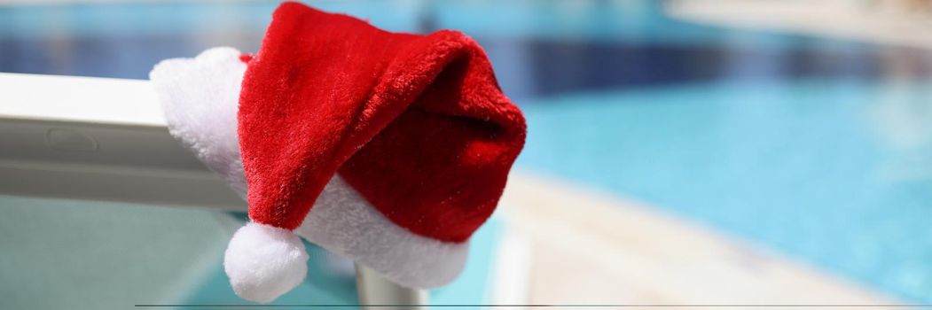 Close-up of santa claus hat put on lawn chair near swimming pool with clean turquoise water. Hotel with decoration for new year celebration. Summer concept