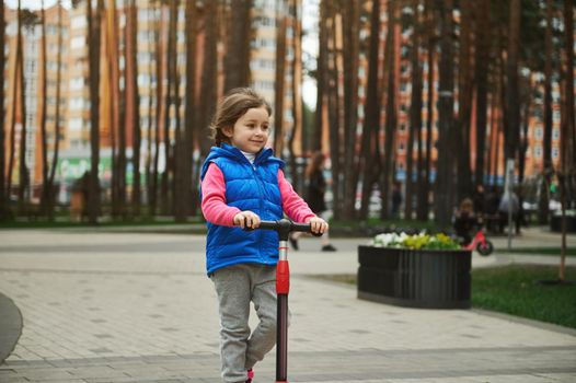Beautiful Caucasian little girl, 5 years old child riding a kick scooter, enjoying sunny day in urban park. Active and healthy lifestyle, happy childhood concept