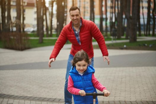 Cheerful father enjoys his day off, spends time with his kid- adorable little girl riding a push scooter in the city park. Happy childhood, fatherhood and family relationships concept