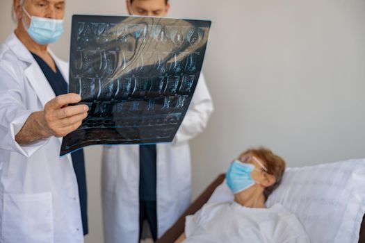 Two doctors in mask look and discuss an X-ray or MRI scan of the patient standing in hospital ward
