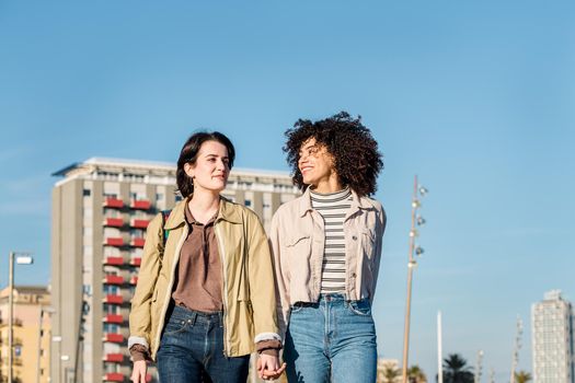 young multiracial couple of girls smiling and walking around the city holding hands, concept of sexual freedom and racial diversity