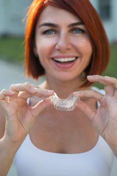 Red-haired Caucasian woman holding transparent mouthguards for bite correction outdoors. A girl with a beautiful snow-white smile uses silicone braces.