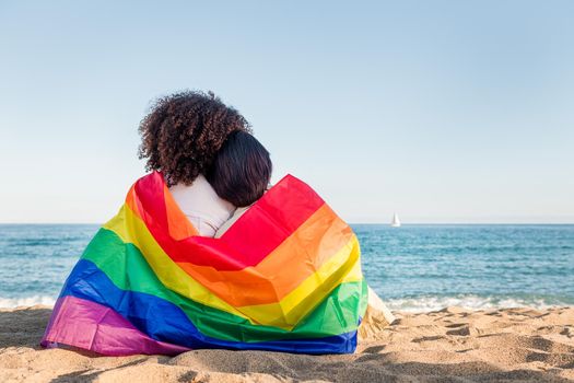 multiracial couple of women in love relaxing by the sea wrapped in the rainbow flag, symbol of the struggle for gay rights, concept of sexual freedom and racial diversity