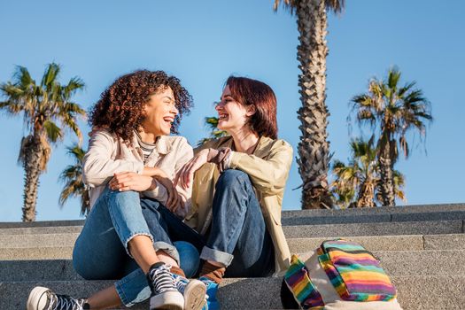 young multiracial couple of homosexual women holding hands and laughing sitting on the stairs, concept of sexual freedom and racial diversity