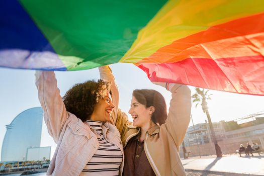 two gay girls laughing with rainbow flag, symbol of the struggle for gay rights, in Barcelona, gay friendly city, concept of sexual freedom and racial diversity, flare effect